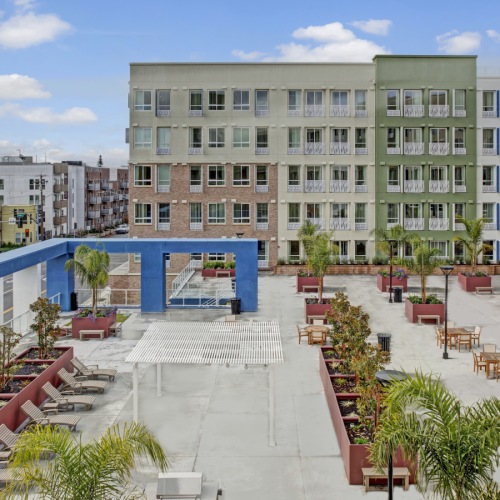 San Jose, CA, Apartments - Virginia Street Studios - Outdoor Courtyard with Brick-Red Plant Boxes, Palm Trees, Lounge Chairs, Covered Areas, and View of Street and Building Exterior in the Background. San Jose, CA, Apartments - Virginia Street Studios - Outdoor Courtyard with Brick-Red Plant Boxes, Palm Trees, Lounge Chairs, Covered Areas, and View of Street and Building Exterior in the Background.