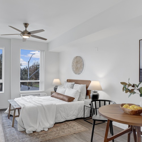 Apartment interior with queen sized bed, table, and chairs Pet-Friendly Apartments in San Jose CA - Virginia Street Studios - A Bright And Spacious Living Area With A Bed Next To Two Very Large Windows With Wood-Style Flooring, A Ceiling Fan, And White Walls.